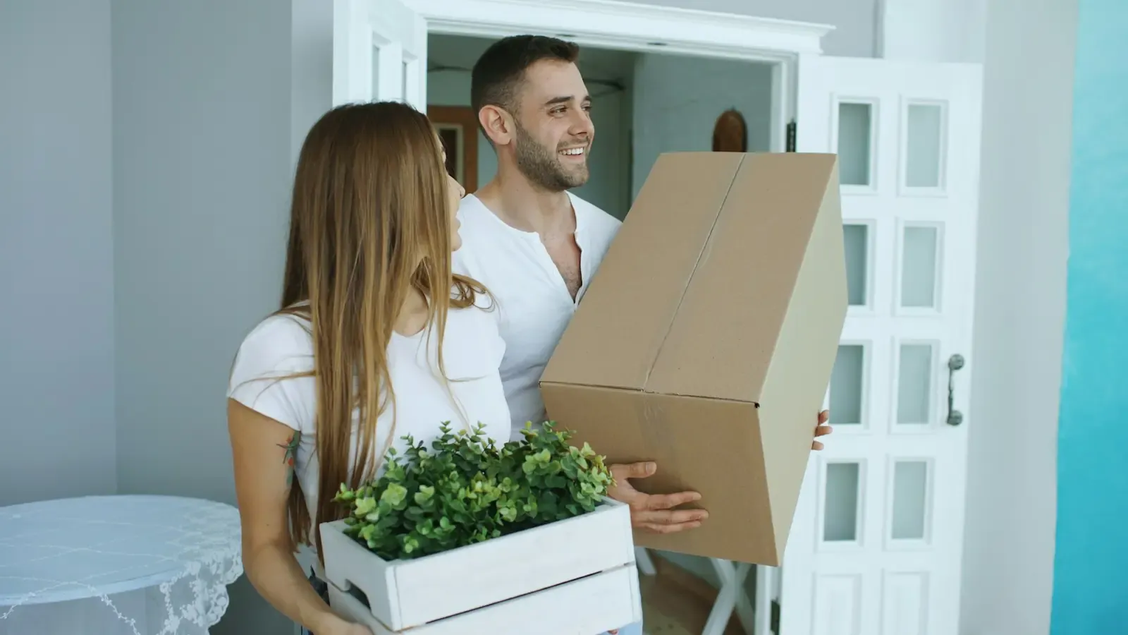 A couple carrying a moving box and a plant into their new home